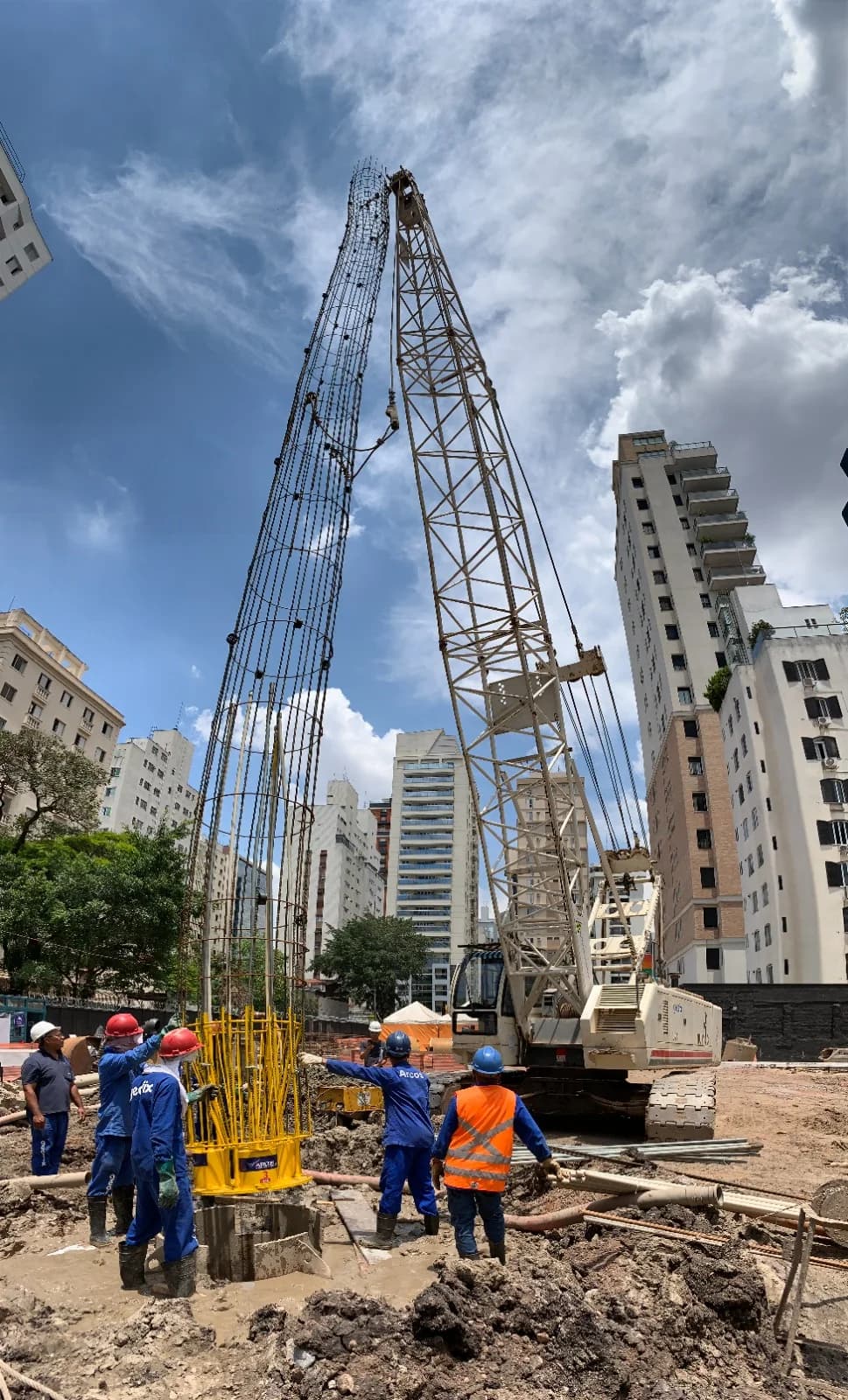 Equipe em canteiro urbano junto à célula de carga bidirecional amarela, com perfuratriz e edifícios ao fundo