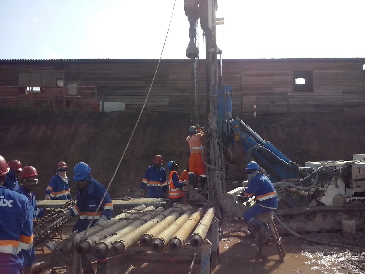 Equipe operando uma perfuratriz de estaca em obra, com hastes de perfuração apoiadas em bancada e armadura de aço sendo manuseada